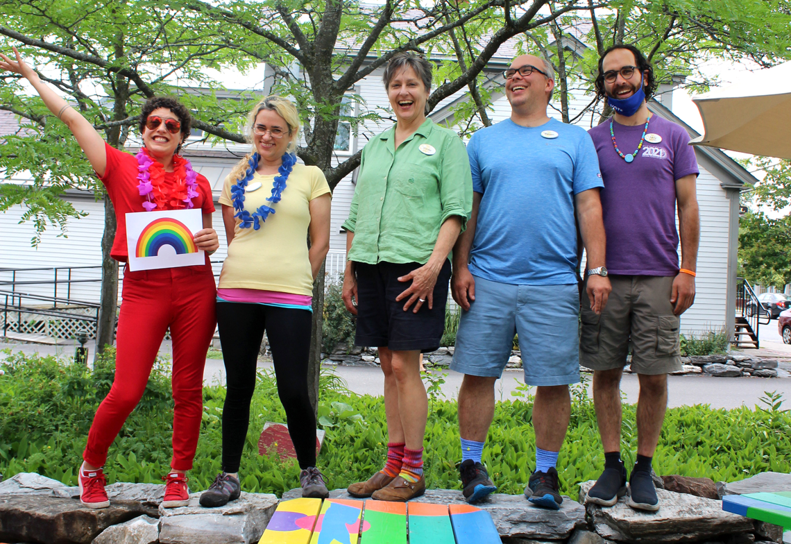 5 co-op employees standing on a rock wall wearing different colored shirts. far left person is wearing a red outfit, holding a rainbow, and looks exuberant. the next employee is wearing a yellow shirt, the person in the middle is wearing a green shirt, the fourth person from left is wearing blue, and the fifth person has a beard and is wearing a purple shirt. All people look happy and proud.